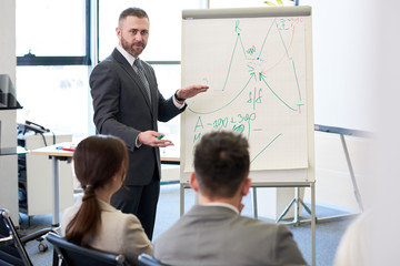 Portrait of bearded business coach standing by whiteboard giving presentation for audience and pointing at statistics graphs