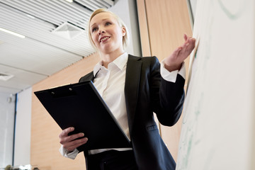 Low angle portrait of blonde businesswoman standing at whiteboard giving presentation to audience