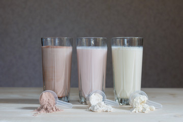 Three milk whey protein shakes in glass cups are arranged in a row with three scoops of protein on a wooden table. Three fresh milk, chocolate, blueberry and banana drinks on table near plastic spoons