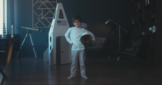 Cute little dreamer boy posing with a helmet at home, pretending to be an astronaut, cardboard space rocket in the background. 4K UHD 60 FPS SLOW MO