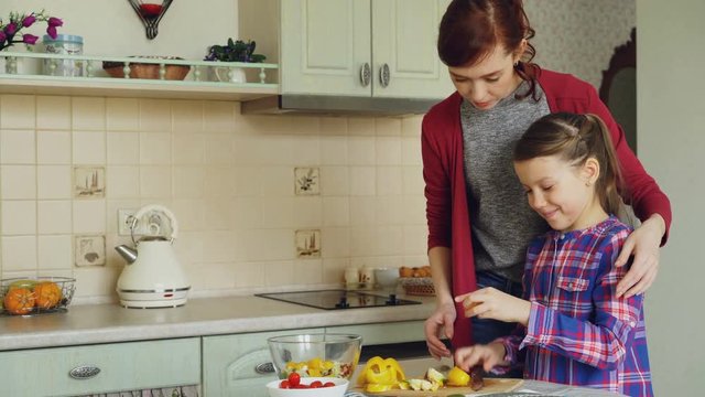 Young pretty mother teaching her cute daughter to cut vegetables properly. Little girl cooking together with loving mom at home in modern kithcen