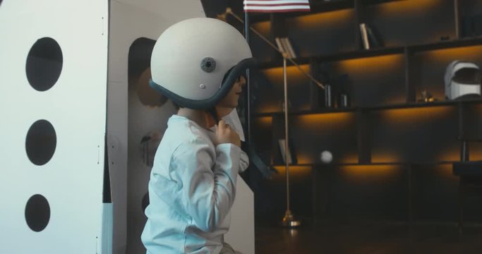 Cute little dreamer boy putting on space helmet at home, pretending to be an astronaut, cardboard space rocket in the background. 4K UHD 60 FPS SLOW MO