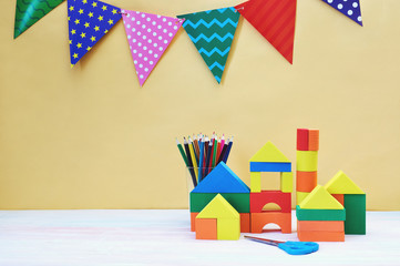 Fototapeta na wymiar Multicolored festive triangular flags, wooden toy house and glass cup with pencils yellow background