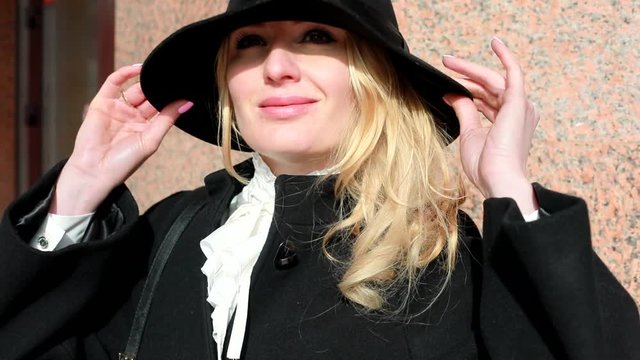 A young woman, a blonde, in a black hat, enjoys the spring sun, is happy to leave for a vacation, a portrait outdoors. The hat is smiling to the camera, talking on the mobile. Against the backdrop of 
