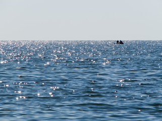 A blue sea with small waves and sun glares, a lonely boat with two men in the distance, and a clear blue sky