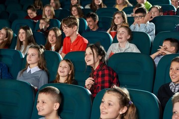 Funny photo of little emotional children watching interesting movie in cinema hall, sitting in comfortable sits,eating popcorn and drinking cola. Kids laughing, smiling, feeling happy and satisfied.