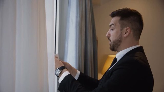Elegant bearded man in formal outfit near big window in hotel and learning speech making marks in keynotes.
