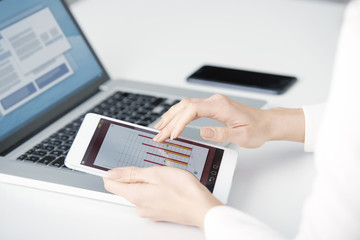 Close-up of businesswoman's hand touching digital tablet and analyzing financial data while sitting in front of laptop at office desk and working on new business report. Isolated on white background. 