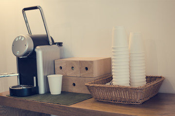  Everyday things - capsule coffee machine stand on wooden table, next to white paper cup folding stack in wicker basket. Toned with warm instagram filters.