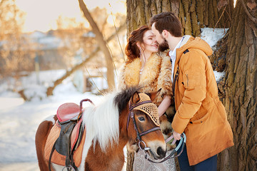 The bride and groom kiss near to a little horse in a winter park at sunset. Winter wedding.