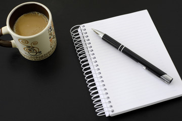 Coffee, notebook and pen on black wooden table. Coffee is an important drink in the workplace.