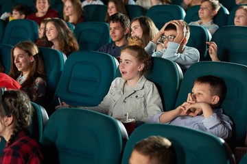 Sideview of laughing children watching movie in the cinema hall. Kids look funny, happy and satisfied. Boys and girls wearing colorful clothes with different prints and smiling.