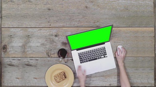 Overhead top view shot of a woman working on a laptop on a wooden desk.  Green screen.  Woman is typing and takes a sip of tea.  Snack and beverage on desk.  Long shot.   HD.