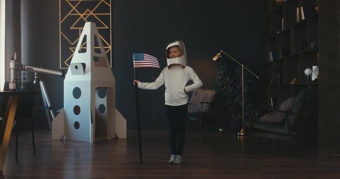 CINEMAGRAPH - seamless loop. Cute little dreamer girl wearing cardboard helmet pretending to be astronaut on Moon, placing US flag near cardboard space rocket at home