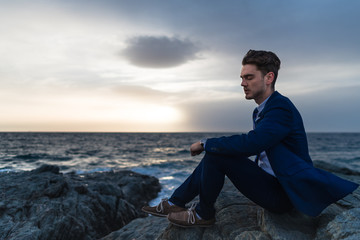 Tired young man in elegant suit sits on the background of the sea and sky. Pensive guy meditating near the ocean in the evening