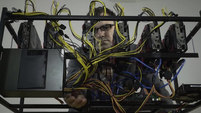 Young adult in work coat maintains a mining rig machine, man checking cable connections of six graphics cards to motherboard obviously, concept cryptocurrency computer, room interior, portrait.
