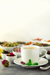 Tea concept with copy space. Different kinds of dry tea in white ceramic bowls and cup of aromatic tea on grey background.