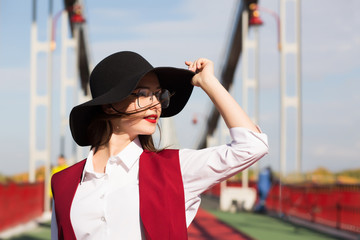 Lifestyle portrait of lovely young model posing at the bridge. Woman wearing fashionable hat, scarf and glasses