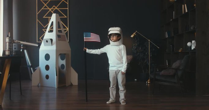 CINEMAGRAPH - seamless loop. Cute little dreamer kid boy wearing cardboard helmet pretending to be astronaut on Moon, placing US flag near cardboard space rocket at home