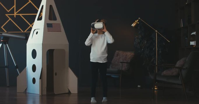 FIXED Little girl wearing VR virtual reality glasses standing near cardboard rocket, studying a model of a space solar system. 4K UHD 