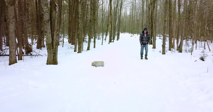 Drone shot of a young man walking in a snowy forest, his dog hangs back on the path behind him.  4K.