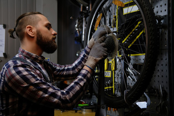Obraz na płótnie Canvas Handsome stylish male wearing a flannel shirt and jeans coverall, working with a bicycle wheel in a repair shop.