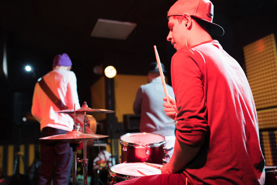 Back view at portrait of modern music band rehearsing in recording studio lit by dim lights while making new album, drummer in foreground