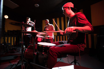 Band of young musicians performing in dark recording studio making new album, drummer in...