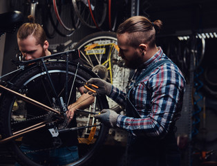 Obraz na płótnie Canvas Two handsome stylish male working with a bicycle in a repair shop. Workers repair and mounts bike in a workshop