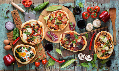 Flatlay of freshly prerared delicious Italian style pizzas. Five pizzas with eggs, salami, mushrooms and vegetables served with raw ingredients and red wine on shabby blue background