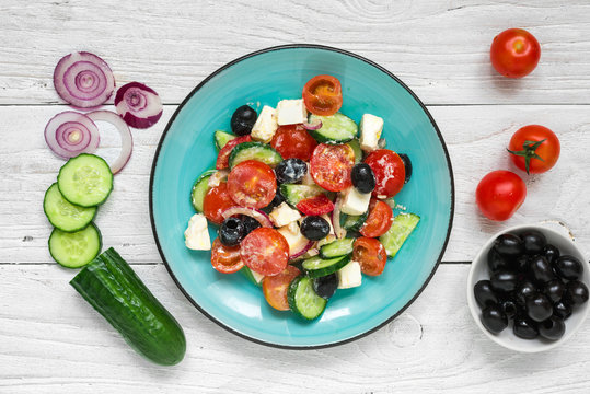 Greek salad with fresh vegetables, feta cheese and black olives in a plate with ingredients on white wooden table