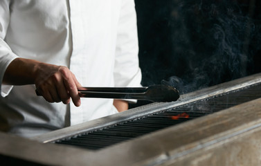 Fototapeta na wymiar Young chef in a white apron standing near the brazier with coals. Man cooking beef steak on barbeque in the interior of modern professional kitchen