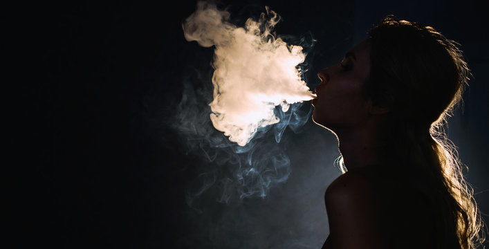 Beautiful young woman profile in backlight smoking hookah with a lot of smoke isolated black background. Club party menu concept