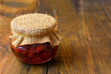 Jar of strawberry jam with a lid made of burlapon dark wooden background