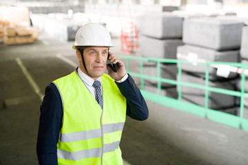 Waist up portrait of mature engineer wearing hardhat and business suit speaking by phone in workshop of modern industrial plant, copy space