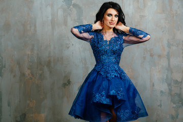 brunette woman in the blue evening dress is posing near the light texture wall background.