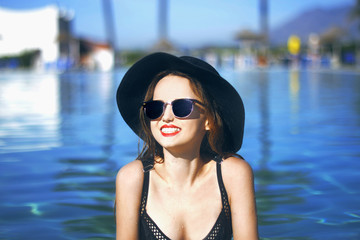 Young beautiful girl in black fashion hat smile, velvet skin, red lips, black swimsuit posing in the pool in blue water, stylish sunglasses, glamor, outdoor portrait, close up, beackground palms.