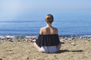 Fototapeta na wymiar Young woman makes meditation in lotus pose on sea / ocean beach, harmony and contemplation. Beautiful girl practicing yoga at sea resort at her vacation. Life style.