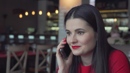 the beautiful and young girl sits at restaurant and speaks by phone. The girl in a red dress speaks by phone and smiles