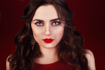 Beautiful luxury woman with jewelry, earrings. Beauty and accessories. Sexy brunette girl with big red lips in a red dress. Fashion model with long curly hair on red background. Smokeys eyes make up.