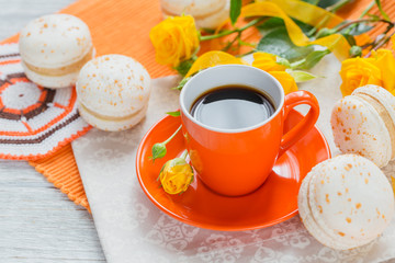 Orange cup of black coffee, yellow roses flowers and sweet pastel french macaroons