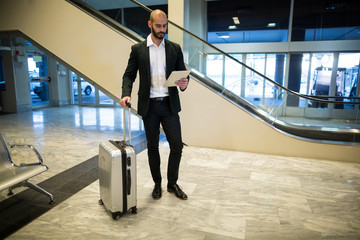 Businessman standing with luggage using digital tablet in waiting area