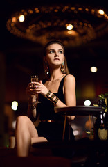 Plakat Beautiful sexy fashion brunette woman in expensive interior restaurant drinking champagne waiting on dark cafe 