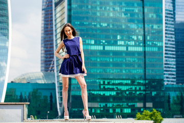 Fototapeta na wymiar Young woman in a blue dress is stretching near skyscrapers