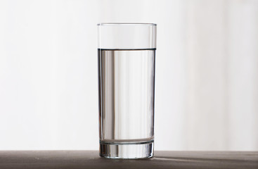 A simple tall glass filled with clear water stands on a wooden surface on a light background. Front...