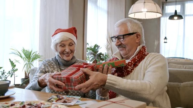 Joyous senior man and woman sitting on couch at restaurant table and sharing Christmas gifts at holiday dinner