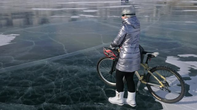 Woman is walking beside bicycle on the ice. The girl is dressed in a silvery down jacket, backpack and helmet. Ice of the frozen Lake Baikal. The tires on the bicycle are covered with special spikes