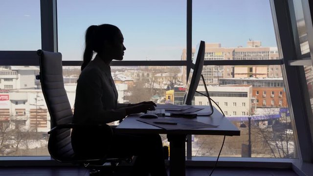Silhouette of business woman who works at computer and talking on phone near window in office. Concept: window in office, business woman, computer and telephone