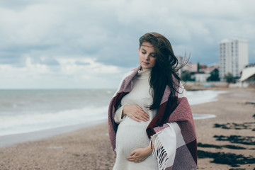 Young beautiful pragnant woman in a white knitted sweater posing near sea at the beach