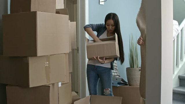 Tilt up of young Asian woman looking for something in pile of cardboard boxes and talking to her husband while unpacking stuff after moving house